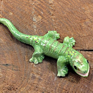 May include: A ceramic lizard figurine in shades of green, with brown and yellow spots, rests on a textured, brown wooden surface. The lizard has a detailed design, including visible claws and a slightly open mouth. The figurine is a decorative item.
