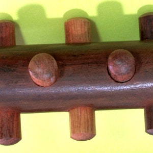 Puede incluir: Un rompecabezas de madera hecho a mano con un cuerpo oblongo marrón oscuro y seis clavijas cilíndricas. Dos clavijas redondeadas están incrustadas en el cuerpo. El rompecabezas es de madera y tiene un acabado liso.