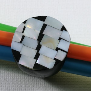 May include: A circular black button featuring a mosaic pattern of white and iridescent rectangular tiles. The button is set against a backdrop of green, orange, and blue cylindrical objects.