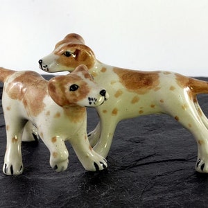 Könnte beinhalten: Zwei Keramik-Hundefiguren. Die Hunde sind weiß mit braunen Flecken und Markierungen auf Kopf und Rücken. Sie sind in einer spielerischen Haltung dargestellt, wobei ein Hund sich an den anderen zu lehnen scheint. Die Figuren stehen auf einer dunklen, strukturierten Oberfläche.