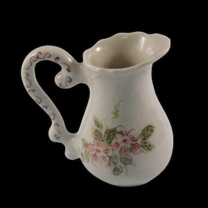 Könnte beinhalten: Ein kleiner, cremefarbener Keramikkrug mit gebogenem Henkel und ausgestelltem Ausguss. Der Krug ist mit rosa und grünen Blumenmustern verziert. Der Griff hat ein dekoratives Wirbelmuster.