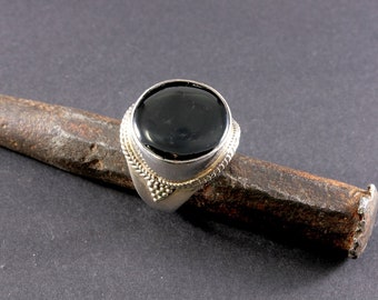 Op de afbeelding: Een zilveren ring met een zwarte edelsteen in een ronde zetting. De ring ligt op een roestig metalen voorwerp.