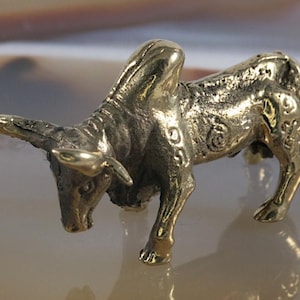 Puede incluir: Una pequeña figura de bronce de color dorado de un toro. El toro está de pie con la cabeza baja y los cuernos apuntando hacia adelante. La figura está detallada con intrincados tallados y patrones.