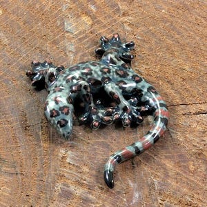 Könnte beinhalten: Keramik-Gecko-Figur mit einem gefleckten Muster in Grau-, Braun- und Rottönen. Der Gecko ist auf einer Holzoberfläche platziert und zeigt sein detailliertes Design und seine strukturierte Oberfläche. Der Schwanz ist elegant geschwungen.