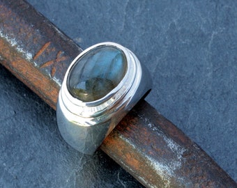 Może przedstawiać: Srebrny pierścionek z dużym owalnym kamieniem labradoritem osadzonym w oprawie typu bezel. Pierścionek ma szeroką obrączkę z fakturowaną powierzchnią.
