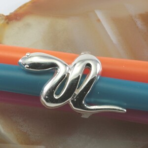Puede incluir: Un anillo de serpiente de plata con un diseño detallado. El anillo está sobre un fondo multicolor.