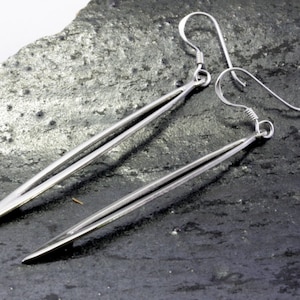 May include: A pair of long, silver earrings with a simple, sleek design. The earrings are made of two thin, parallel lines that taper to a point at the bottom.