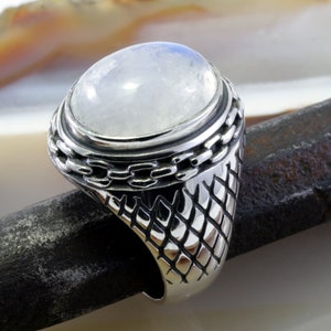 Puede incluir: Un anillo de plata con una gran piedra de luna ovalada engastada en una banda texturizada. La banda tiene un diseño de cadena alrededor de la piedra.