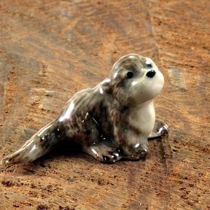 Könnte beinhalten: Eine kleine Keramikfigur eines Otters mit glänzender Oberfläche. Der Otter ist in Braun-, Grau- und Weißtönen gehalten und zeichnet sich durch einen glatten, abgerundeten Körper, einen spitzen Schwanz und ein kleines Gesicht mit schwarzen Augen und Nase aus.