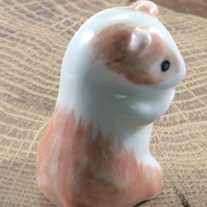 May include: A ceramic guinea pig figurine. The figurine is white with patches of reddish-brown on its head and body. It has a small black eye and is posed in a seated position. The figurine is on a wooden surface.