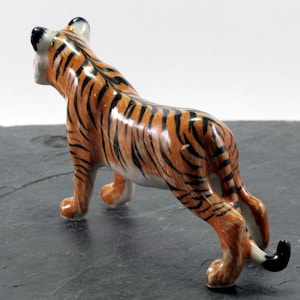 Op de afbeelding: Een keramieken tijgerfiguur met oranje en zwarte strepen. De tijger staat in een loopstand, met zijn staart omhoog. Het beeldje heeft een glanzende afwerking en staat tegen een donkere achtergrond.