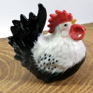 Könnte beinhalten: Eine weiße und schwarze Keramik-Hahnfigur mit rotem Kamm und Kehllappen. Der Hahn sitzt auf einer Holzoberfläche.