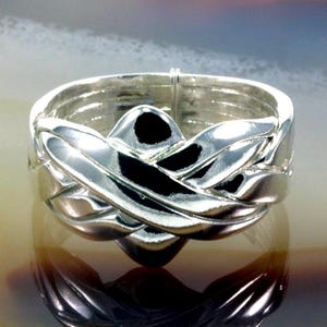 May include: A silver puzzle ring with an intricate, interwoven design. The ring's bands are smooth and reflective, creating a polished appearance. The puzzle ring is a unique piece of jewellery.
