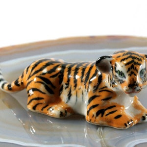 Könnte beinhalten: Eine Keramik-Tigerfigur in Ruheposition. Der Tiger hat orange und schwarze Streifen, mit weißem Bauch und Gesicht. Die Figur steht auf einer hellen, gemusterten Oberfläche.