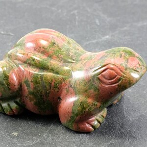 Könnte beinhalten: Eine geschnitzte Froschfigur aus Stein in Rot- und Grüntönen. Der Frosch ist mit geschnitzten Augen, Beinen und einem strukturierten Körper detailliert. Die Figur besteht aus poliertem Stein, und die Farben sind natürlich.