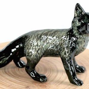 Könnte beinhalten: Keramikfuchsfigur mit glänzendem Finish. Der Fuchs ist überwiegend schwarz und grau mit einer weiß getippten Schwanzspitze. Er ist in einer Laufhaltung auf einer Holzoberfläche positioniert und zeigt seine detaillierten Merkmale und Handwerkskunst.
