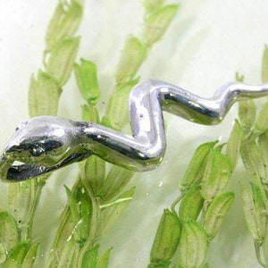 Pode incluir: Uma figura de cobra prateada e estilizada, com um corpo curvo e uma cabeça detalhada, é colocada entre plantas aquáticas verdes. A cobra parece estar nadando na água, criando uma cena dinâmica e naturalista.