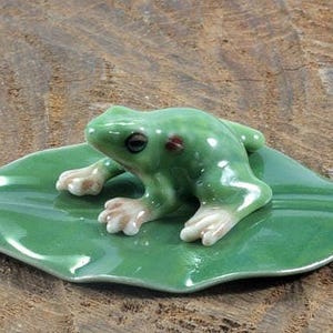 Könnte beinhalten: Eine glänzende, grüne Keramikfroschfigur, die auf einem grünen Blatt sitzt. Der Frosch hat weiße Füße und ein detailliertes Gesicht. Das Blatt ist gebogen. Die Figur steht auf einer Holzoberfläche.