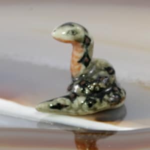 Könnte beinhalten: Kleine Keramik-Schlangenskulptur mit gewundenem Körper und aufrechtem Kopf. Die Schlange hat ein hellgrünes und schwarzes, gesprenkeltes Muster mit orangefarbenen und braunen Akzenten. Die Figur steht auf einer reflektierenden Oberfläche.