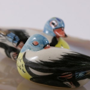May include: Two ceramic duck figurines, one blue and white with black accents, the other black and white with yellow accents. The ducks are lying down with their heads turned to the side.