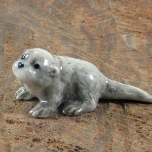 May include: A small, ceramic otter figurine in shades of grey. The otter is depicted in a crawling position with its head raised, showcasing detailed features. The figurine is placed on a textured, brown surface.