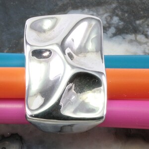 May include: A polished silver ring with a rectangular shape and an uneven, textured surface. The ring has an organic design. The background features blue, orange, and pink pencils.
