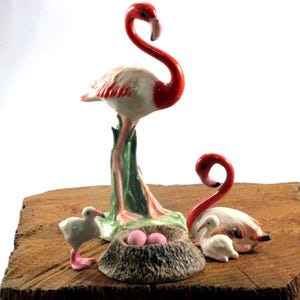 Op de afbeelding: Een keramisch flamingo familie beeldjes set. De set bevat een grote flamingo met een rode nek en witte lichaam, een nest met roze eieren, twee kleinere flamingo's en een baby flamingo. De beeldjes staan op een houten oppervlak.