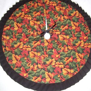 May include: Circular tree skirt with a fall leaf pattern in shades of red, orange, and green. The skirt has a black pleated border and a black ribbon tie. A festive decoration for the holiday season.