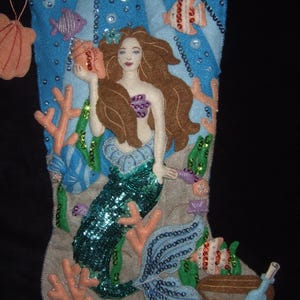 May include: A handmade Christmas stocking featuring a mermaid design. The mermaid has brown hair and a sequined teal tail, holding a seashell. The stocking is decorated with coral, fish, and sequins, with a treasure chest at the bottom.