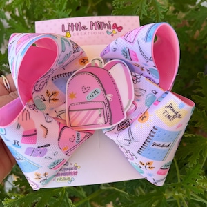 May include: A large, pink and white hair bow with a school-themed design. The bow features a pink backpack with the word "CUTE" on it. The bow also has a pattern of school supplies and the words "Study Time" and "Notebook". The bow is made of ribbon.