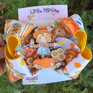 May include: A large hair bow featuring a white ribbon with a repeating autumn pattern of scarecrows, sunflowers, pumpkins, and blue birds. A central layered embellishment depicts a scarecrow with pumpkins. The card reads Little Mimi Creations.