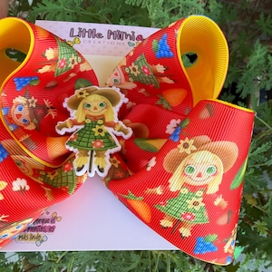 May include: A large red hair bow made of ribbed fabric featuring a repeating pattern of scarecrows, sunflowers, and blue berries. A central scarecrow charm is attached to the front. The bow is mounted on a white card labeled Little Mimi Creations.
