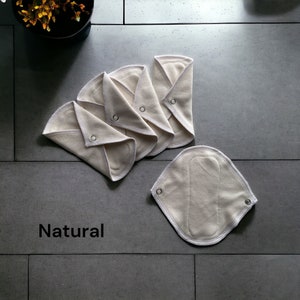 K&ouml;nnte beinhalten: F&uuml;nf wiederverwendbare Stoffbinden in Naturfarbe mit wei&szlig;en N&auml;hten und Metalldruckkn&ouml;pfen. Die Binden sind gefaltet und in einem Stapel auf einer grauen Fliesenfl&auml;che angeordnet. Das Wort "Natur" ist in Schwarz unter den Binden gedruckt.