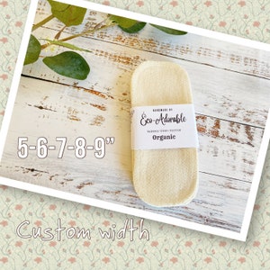 Puede incluir: Un primer plano de una compresa menstrual reutilizable de bambú, cáñamo y algodón orgánico de color blanco. La compresa está etiquetada como "Eco-Adorable" y "Orgánica". La compresa tiene 5-6-7-8-9 pulgadas de largo. El texto "Ancho personalizado" está escrito debajo de la compresa.