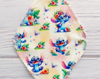 May include: A reusable cloth menstrual pad featuring a repeating pattern of the blue alien character Stitch from Lilo & Stitch. The design includes Stitch with flowers, butterflies, and tropical foliage on a cream background. The pad has a colorful stitched edge and snap closure.