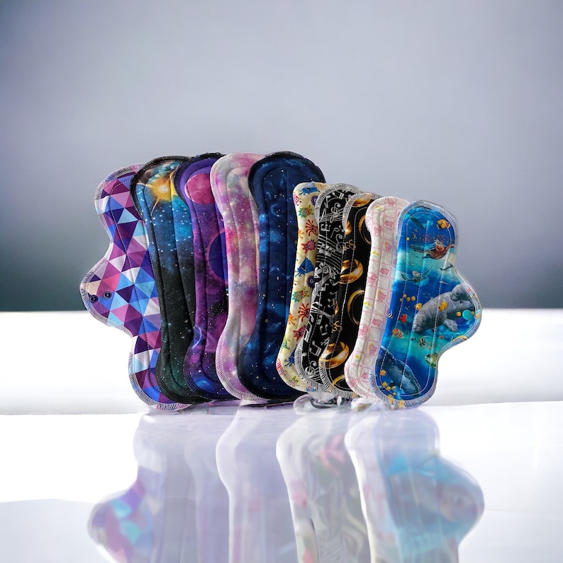 May include: A set of reusable cloth menstrual pads in various patterns, including galaxy prints, musical notes, and a sea creature design.