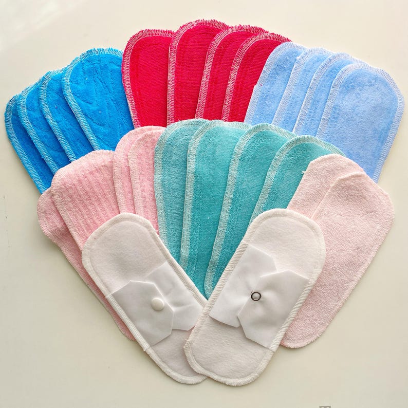 K&ouml;nnte beinhalten: Eine Sammlung wiederverwendbarer Stoffbinden in verschiedenen Farben, darunter Blau, Rot, Rosa und T&uuml;rkis. Die Binden haben eine weiche, saugf&auml;hige Textur und sind mit einer wei&szlig;en R&uuml;ckseite mit Knopfverschluss versehen.