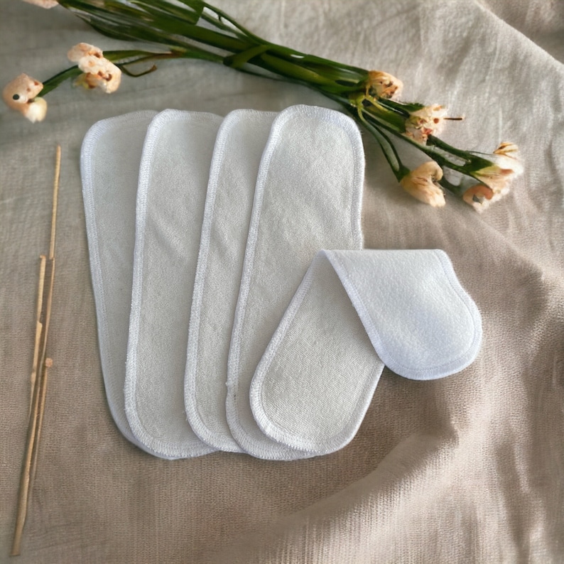 May include: A set of five white, rectangular, absorbent fabric inserts with rounded corners and finished edges. These soft, multi-layered cloth pads are designed for use as diaper liners, featuring a consistent texture and clean, white appearance.