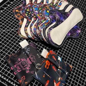 May include: A stack of reusable cloth menstrual pads with various colorful and patterned designs. The pads are folded and arranged on a black and white grid surface. Two small fabric pouches with different designs are also visible.