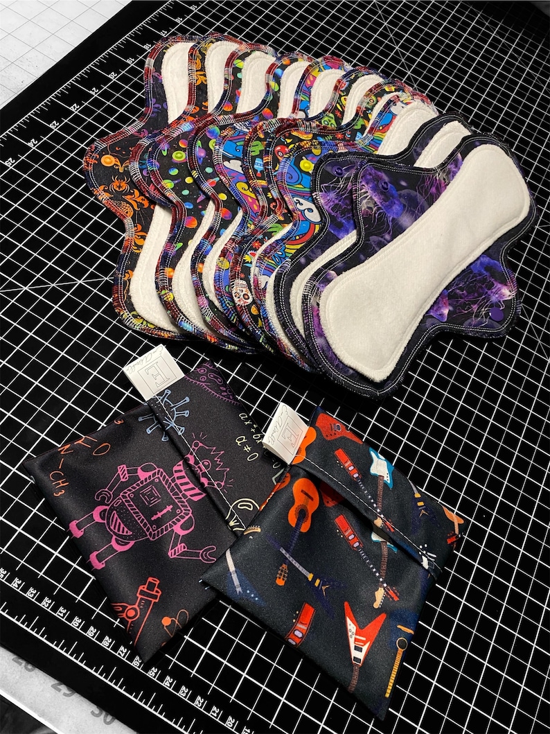 May include: A stack of reusable cloth menstrual pads with various colorful and patterned designs. The pads are folded and arranged on a black and white grid surface. Two small fabric pouches with different designs are also visible.