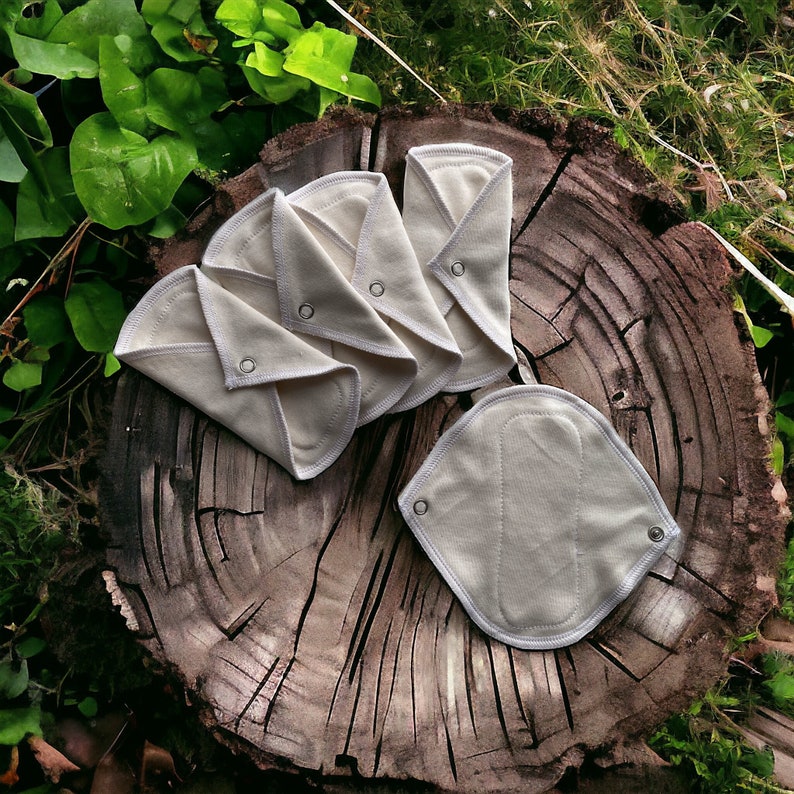 K&ouml;nnte beinhalten: F&uuml;nf wei&szlig;e, wiederverwendbare Menstruationseinlagen aus Baumwolle, die in der H&auml;lfte gefaltet sind, und eine einzelne Einlage, die flach auf einem Baumstumpf liegt. Die Einlagen bestehen aus wei&szlig;em Baumwollstoff mit wei&szlig;em Besatz und einem silbernen Druckknopf an jeder Seite.
