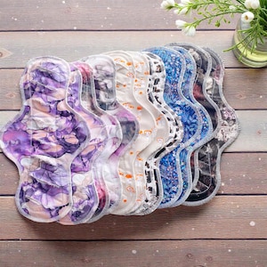 Può includere: Una collezione di assorbenti in tessuto riutilizzabili con vari motivi. Gli assorbenti presentano disegni colorati, tra cui motivi viola e bianchi in stile acquerello, blu floreali e in bianco e nero. Ogni assorbente ha una forma curva e cuciture bianche.