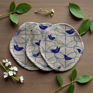 May include: Three round, beige fabric pads with a blue bird and leaf pattern. The pads have a stitched edge and are arranged on a wooden surface, surrounded by green leaves and white flowers.