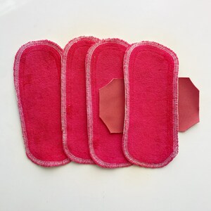 K&ouml;nnte beinhalten: Vier leuchtend rosa, rechteckige Stoffeinlagen mit abgerundeten Kanten und wei&szlig;en N&auml;hten. Ein separates, kleineres, achteckiges St&uuml;ck passenden rosa Stoffes ist ebenfalls vorhanden. Die Einlagen sind auf einer wei&szlig;en Oberfl&auml;che angeordnet.
