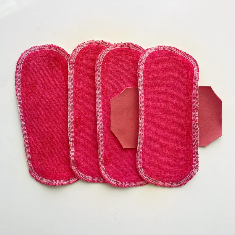 K&ouml;nnte beinhalten: Vier leuchtend rosa, rechteckige Stoffeinlagen mit abgerundeten Kanten und wei&szlig;en N&auml;hten. Ein separates, kleineres, achteckiges St&uuml;ck passenden rosa Stoffes ist ebenfalls vorhanden. Die Einlagen sind auf einer wei&szlig;en Oberfl&auml;che angeordnet.