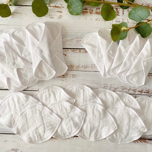 May include: A set of white reusable cloth menstrual pads. The pads are folded and arranged in three groups on a white wooden surface. The pads are made of soft, absorbent fabric and are designed to be comfortable and discreet.