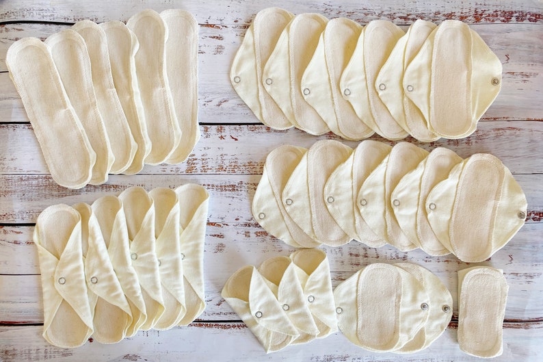 K&ouml;nnte beinhalten: Eine Sammlung wiederverwendbarer Stoff-Menstruationseinlagen in verschiedenen Gr&ouml;&szlig;en und Formen. Die Einlagen bestehen aus nat&uuml;rlicher, ungef&auml;rbter Baumwolle und haben eine hellbeige Farbe. Sie sind auf einer wei&szlig;en Holzoberfl&auml;che angeordnet.