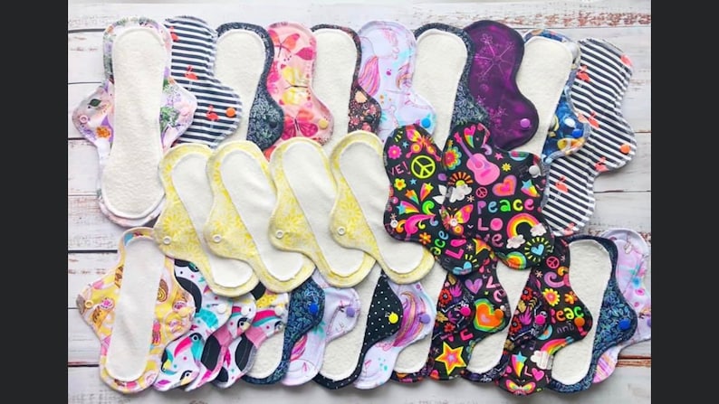 May include: A collection of reusable cloth menstrual pads with various colorful patterns, including floral, geometric, and abstract designs. Some pads feature text like "Peace" and "Love".