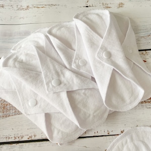 May include: Four white reusable cloth menstrual pads with snap closures. The pads are folded and stacked on a white wooden surface.