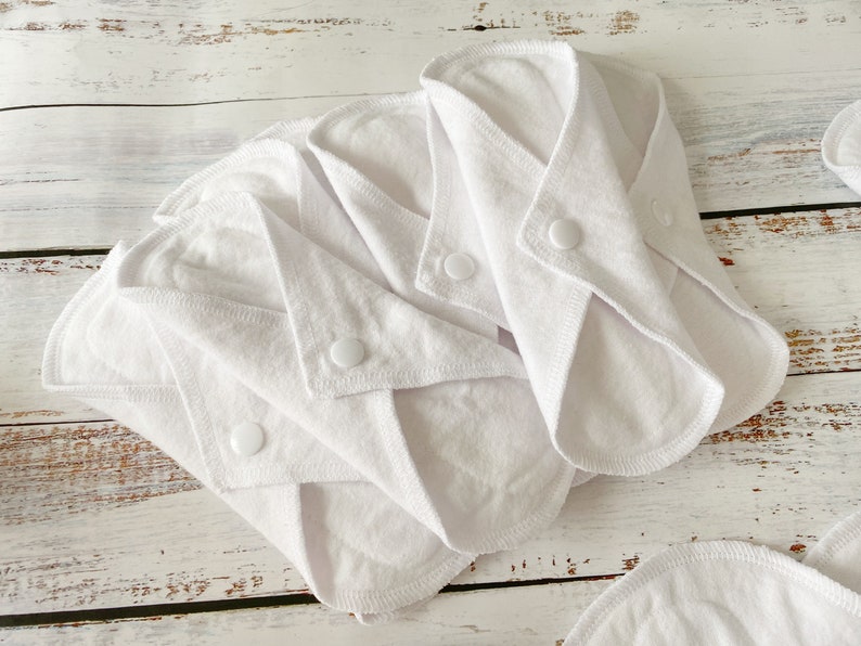 May include: Four white reusable cloth menstrual pads with snap closures. The pads are folded and stacked on a white wooden surface.